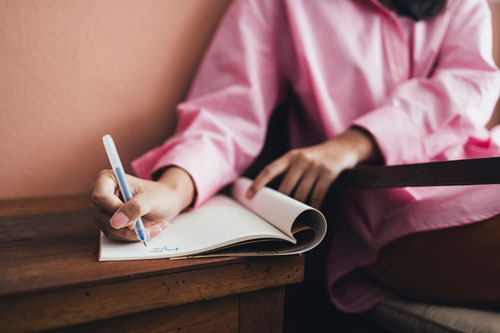 Woman Wearing a Pink Shirt and Orange Trousers Sitting in her Living Room and Writing in her journal