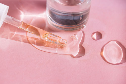 Close-up of drops of facial serum or essential oil with pipette and glass bottle on pink background