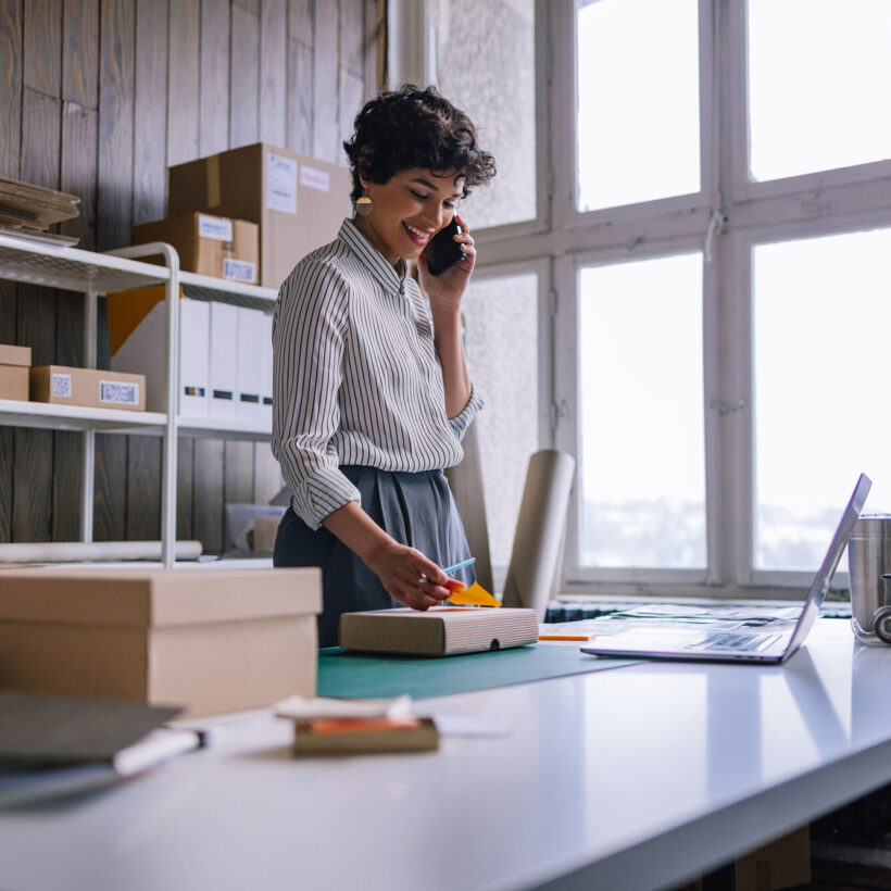 Businesswoman Talking On Her Smartphone While Preparing Packages For Shipping In Her Store