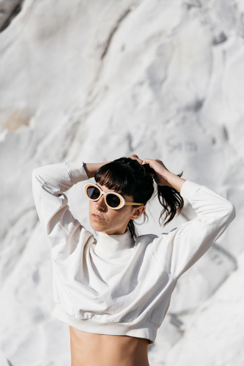 Woman wearing sunglasses tying hair while standing against snow covered rock during sunny day