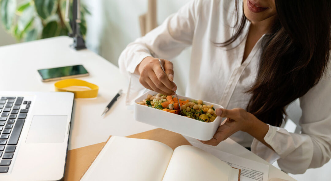 woman eating salad at her desk with laptop and notebook