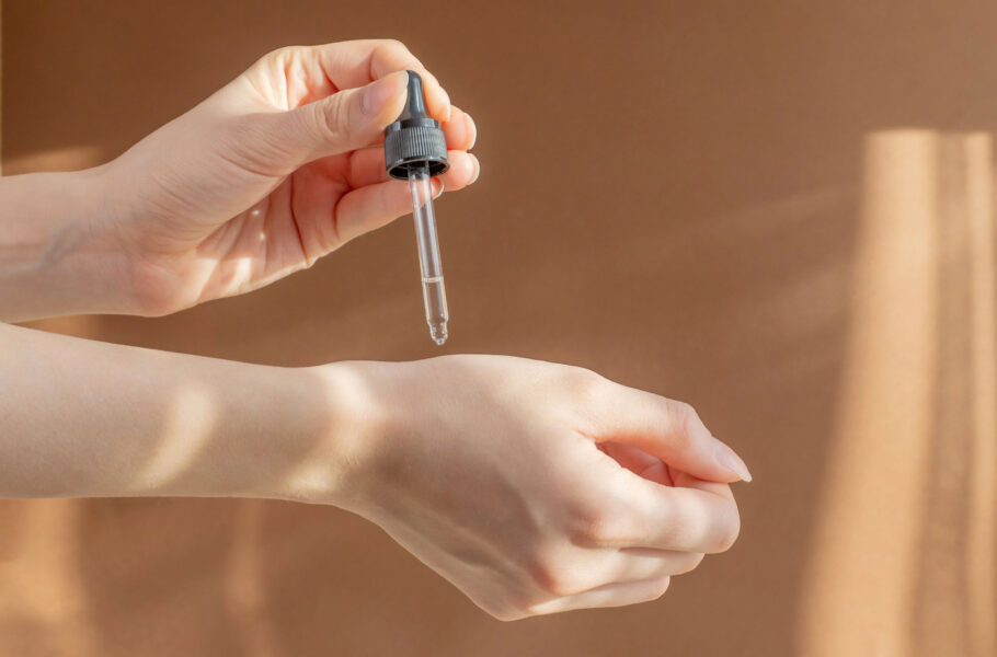 Pipette, with cosmetic serum, in women's hands on light brown background, in rays of sunlight
