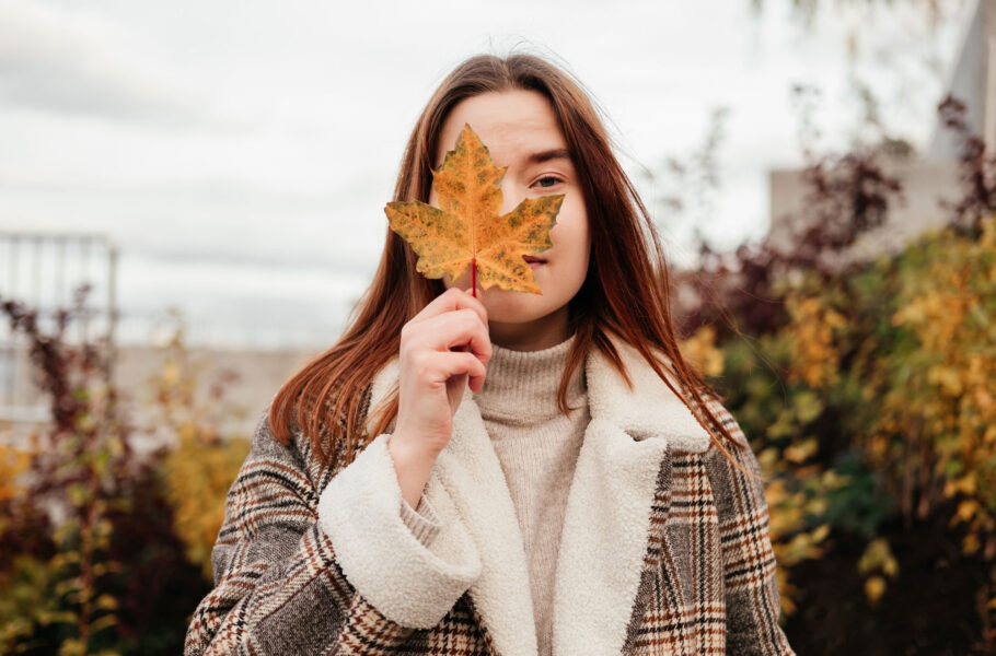woman in coat and sweater holding in hand yellow maple leaf near face and cover one eye