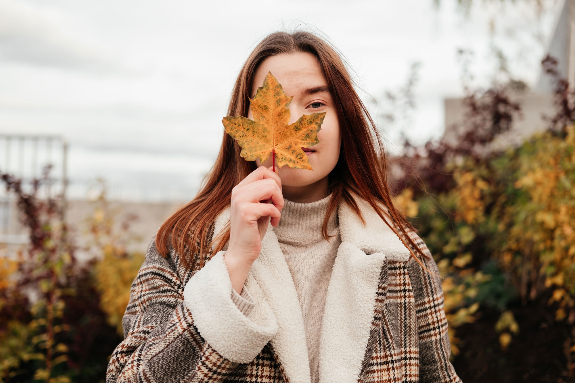 woman in coat and sweater holding in hand yellow maple leaf near face and cover one eye