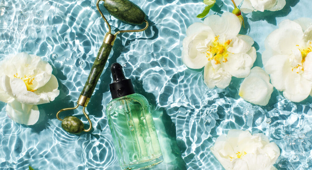 Glass bottle of natural cosmetic serum with a pipette, massage essential oil and Jade face roller. with white rose flowers on a background of blue water with waves