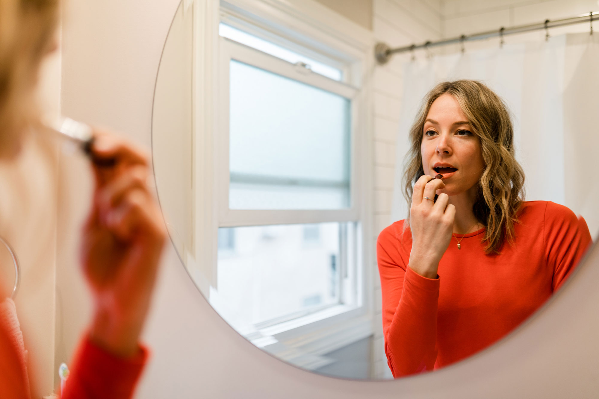 Woman Wearing Red Sweater Puts on Lipstick in Bathroom Mirror