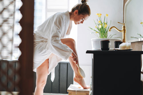 Woman In White Bathrobe Doing Body Massage With Dry Wooden Brush In Bathroom At Home