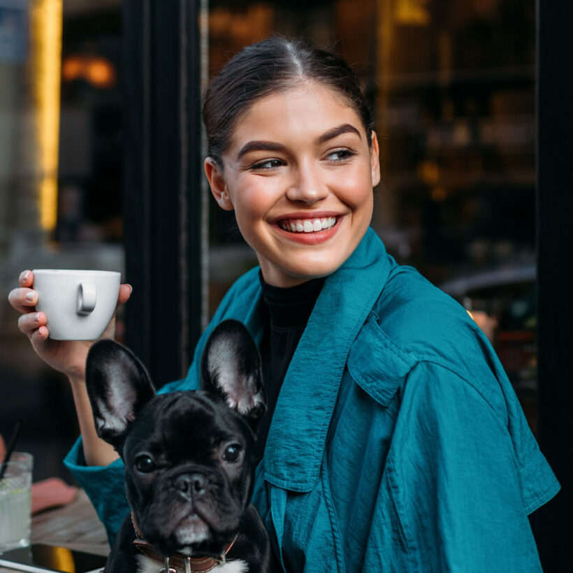 woman with her french bulldog, sitting in front of the restaurant, holding espresso cup and waiting for someone.