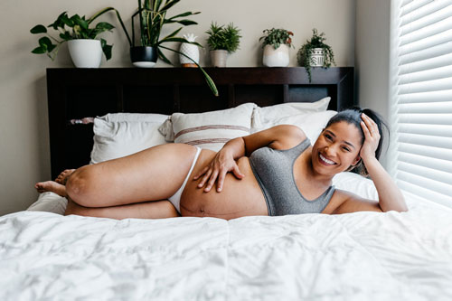 woman laying down in her bed in a bra & panties holding her pregnant belly