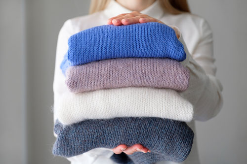 woman holds an armful of knitted things of different colors, stacked in a pile, in the room.
